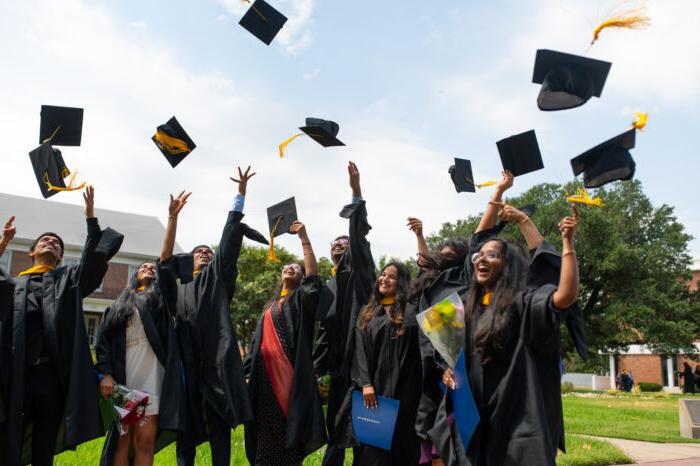 Graduates in black gowns toss their caps into the air. Blue sky with clouds and green grass are in the background.